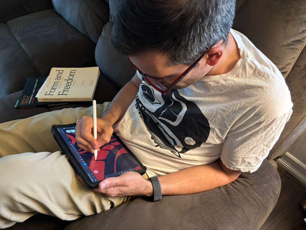 Artist seated on a couch drawing red formline on an iPad. The book Form and Freedom: Northwest Coast Indian Art is open beside him on the cushion.