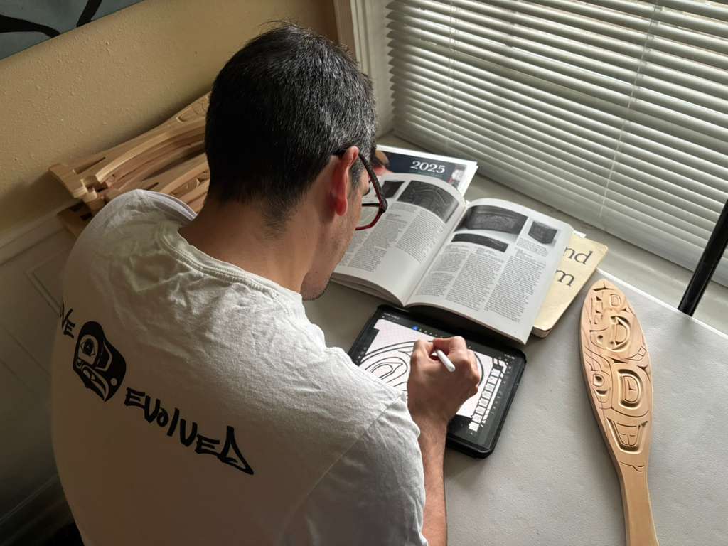 Artist at a desk by a window, drawing on an iPad with reference books open in front of him. A carved wooden paddle with formline design rests on the desk to his right.