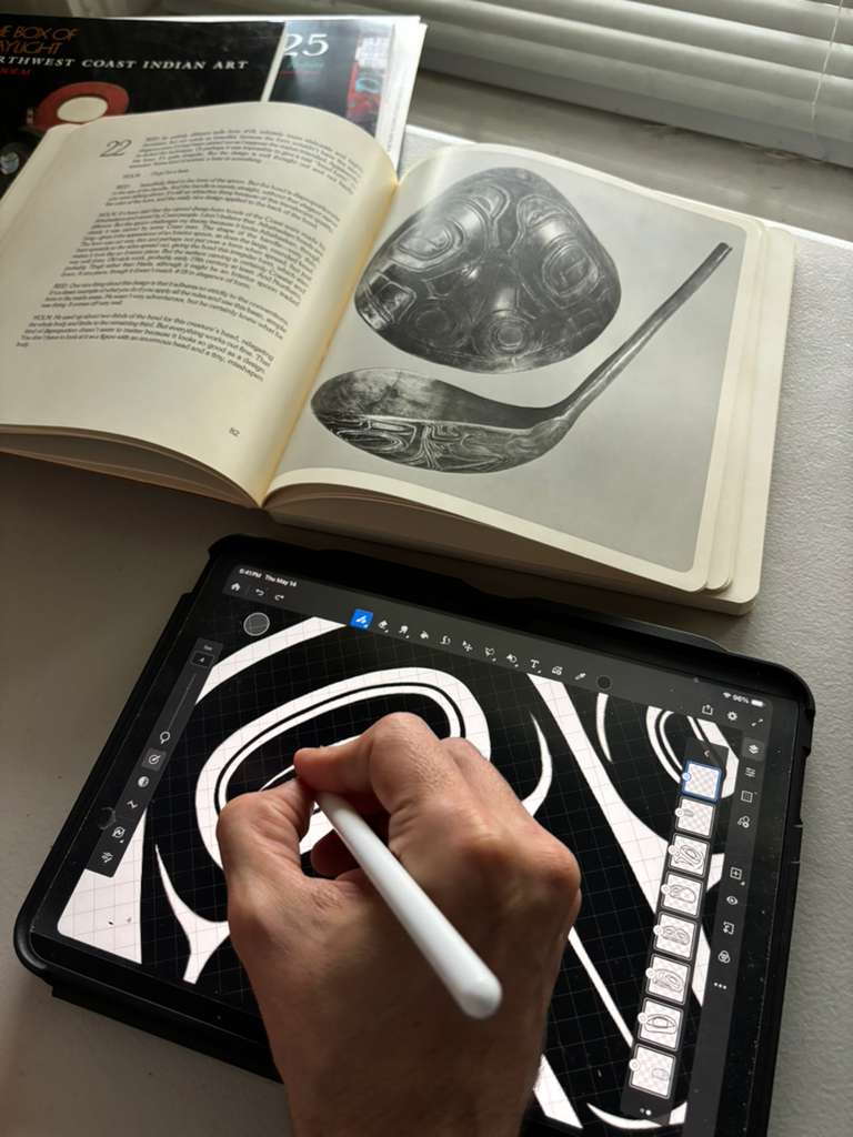Artist bent close over an iPad drawing black and white formline linework with an Apple Pencil. Behind the iPad, a Bill Holm reference book is open to a page of Northwest Coast art analysis.