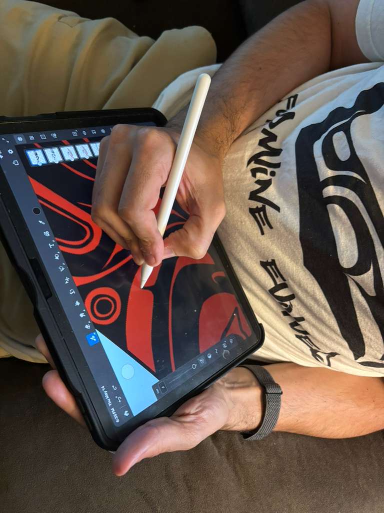 Close up of hands holding an iPad and Apple Pencil, drawing bold black formline shapes against a red background. The Formline Evolved shirt is visible in the frame.