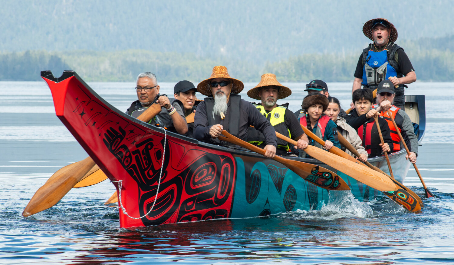 A full crew of paddlers, elders and youth together, paddling the Ancestral Echoes canoe across calm water with forested mountains in the background. The red and teal formline design is vivid against the grey-blue water.