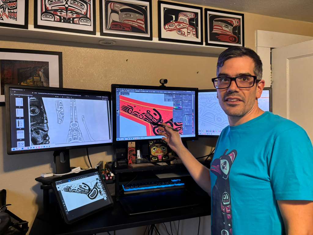 Artist at a triple-monitor workstation with archival reference images on one screen, the canoe template on the second, and the colored design emerging on the third. An iPad with formline sketches sits on the desk.