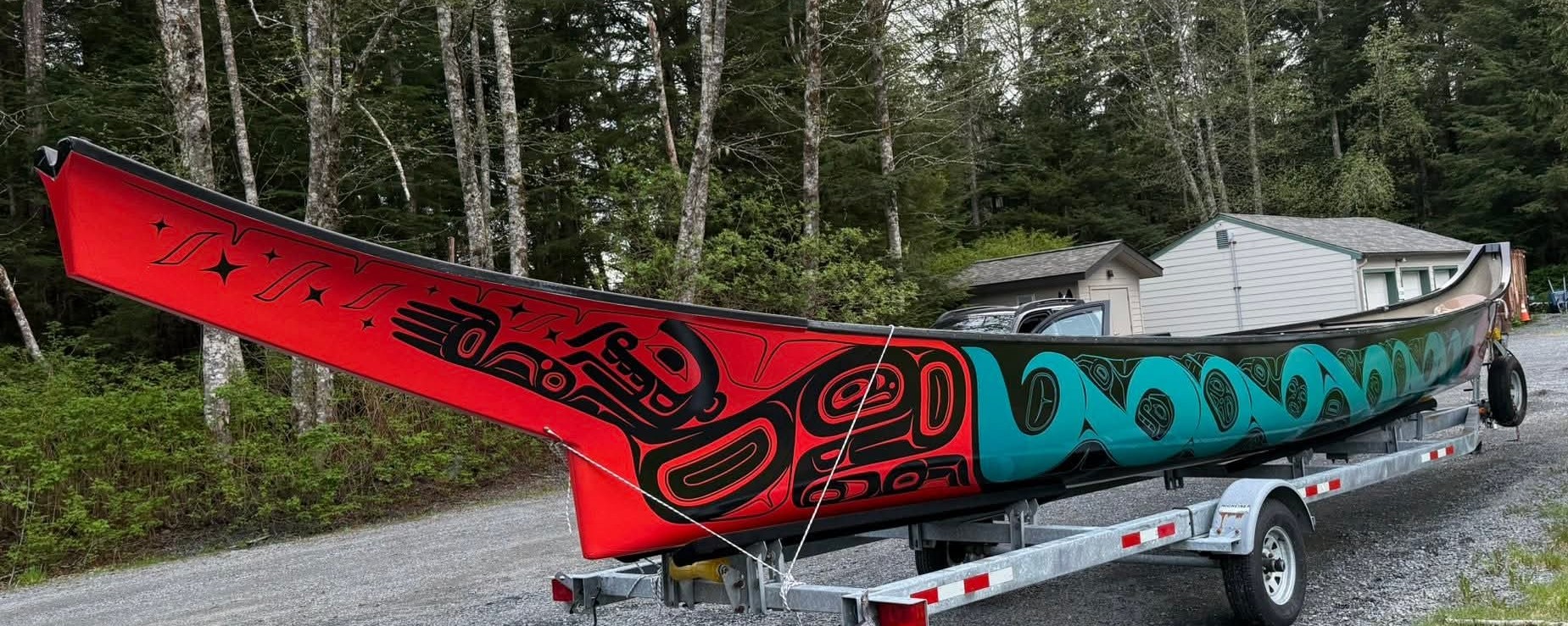The completed canoe on a trailer in a Pacific Northwest forest setting. The bow is in the foreground showing the black figure on red background, stars visible above the figure, transitioning into the teal mid-section toward the stern.