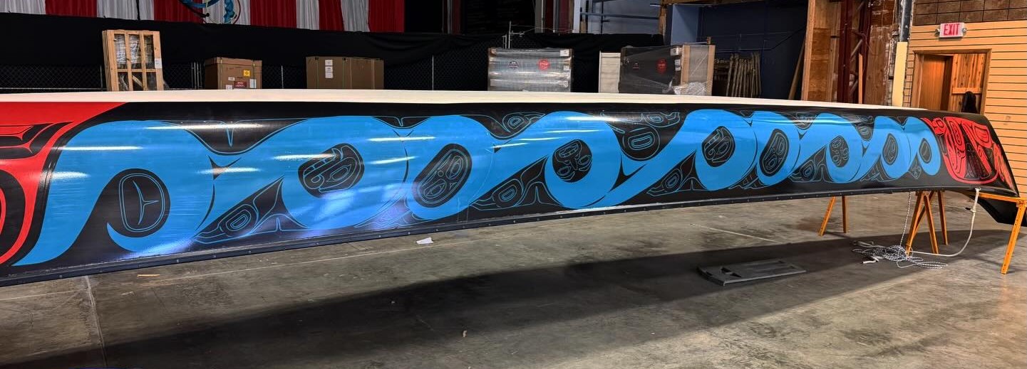 Wide shot of the completed 40-foot canoe wrap displayed upside down in a large warehouse. The full design is visible: the red bow figure on the left, bold teal waves across the mid-section, and the red stern figure on the right against black.